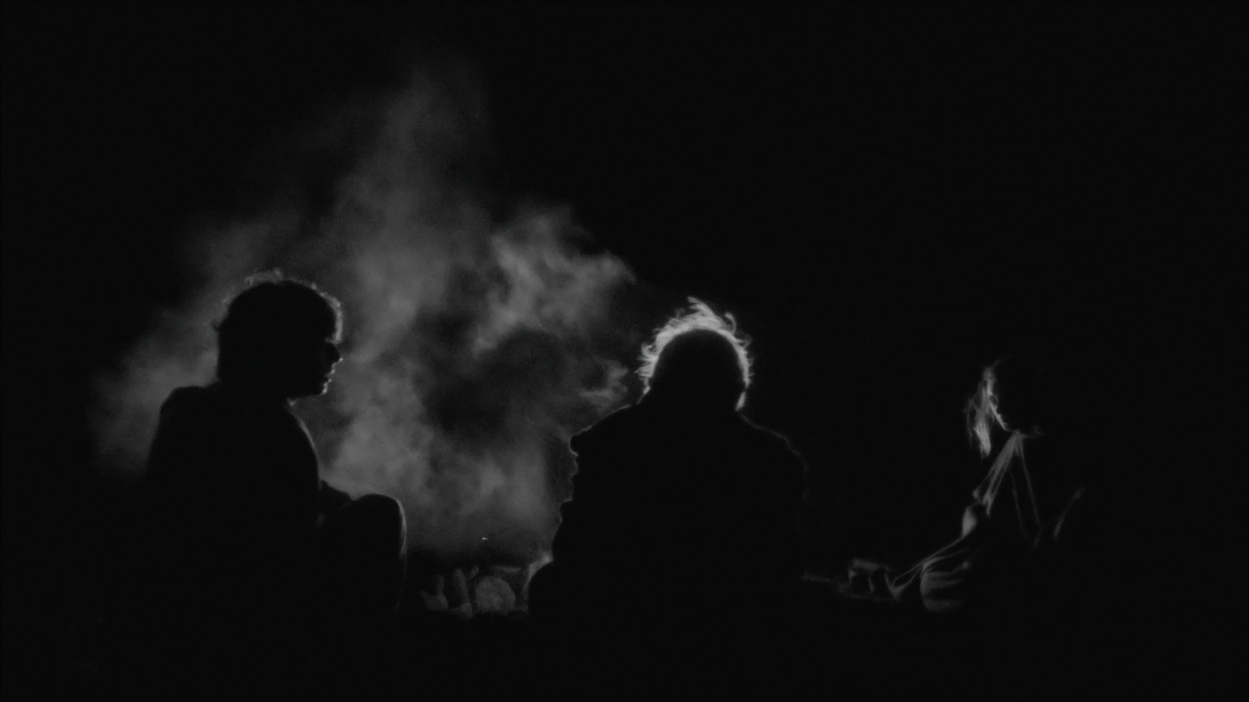 Black-and-white night shot of three people sitting around a campfire in silhouette.