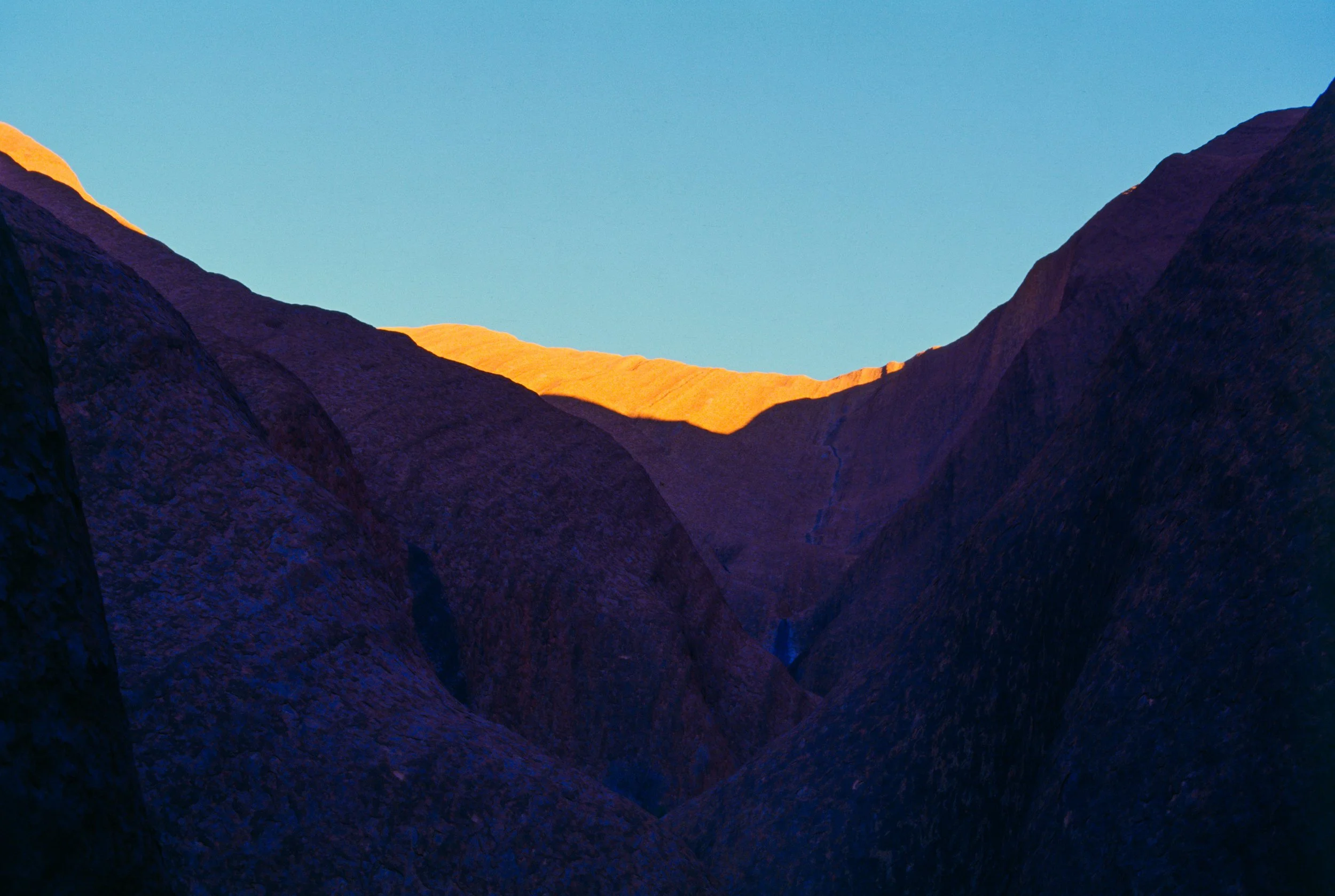 Steep rock walls of Uluru in deep shadow with sunlit sandstone glowing along the ridge