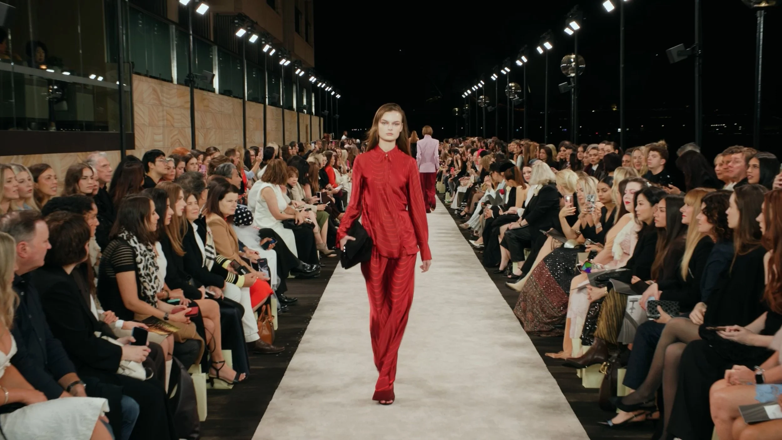 Deep perspective runway shot of a model in a red jumpsuit at a nighttime Sydney fashion show.