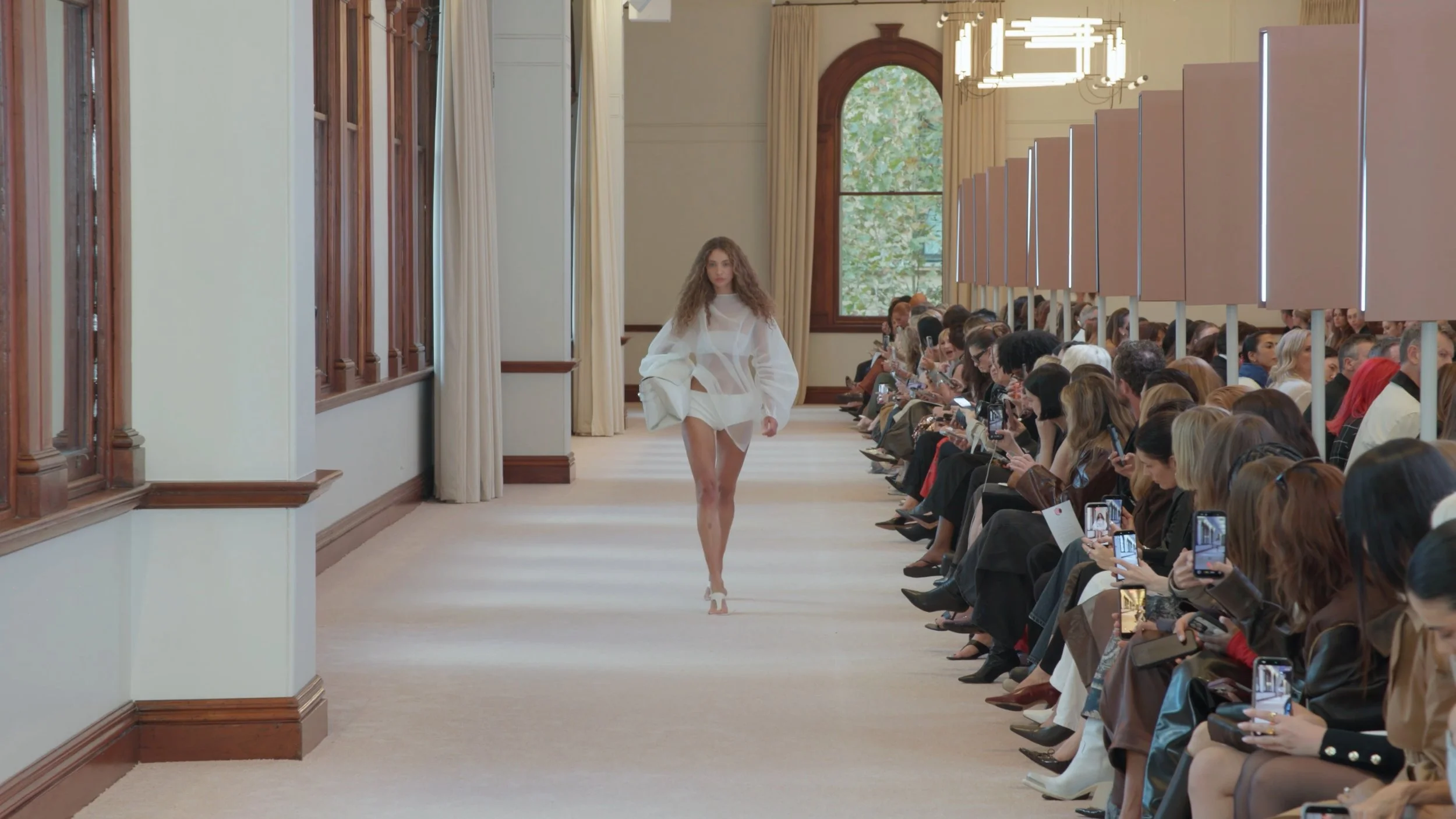 Wide angle runway cinematography of a model in a white avant-garde dress at an AFW Sydney fashion show.