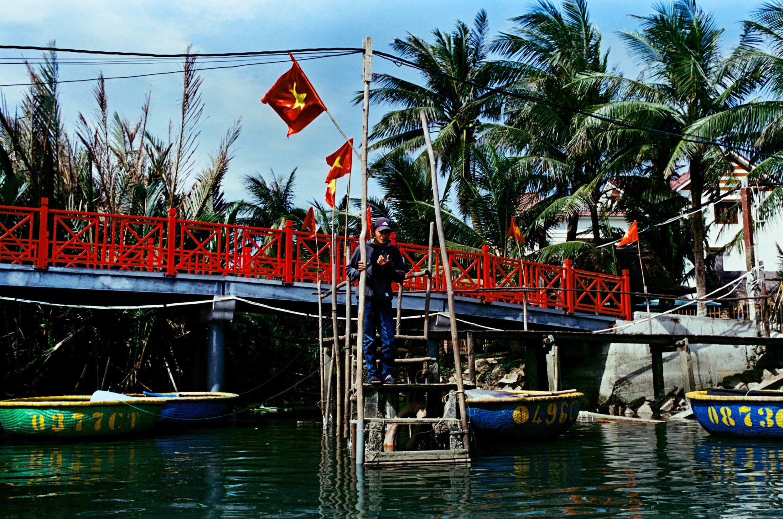 Man standing on a wooden ladder above a river with boats, palm trees, and a red pedestrian bridge in a tropical town