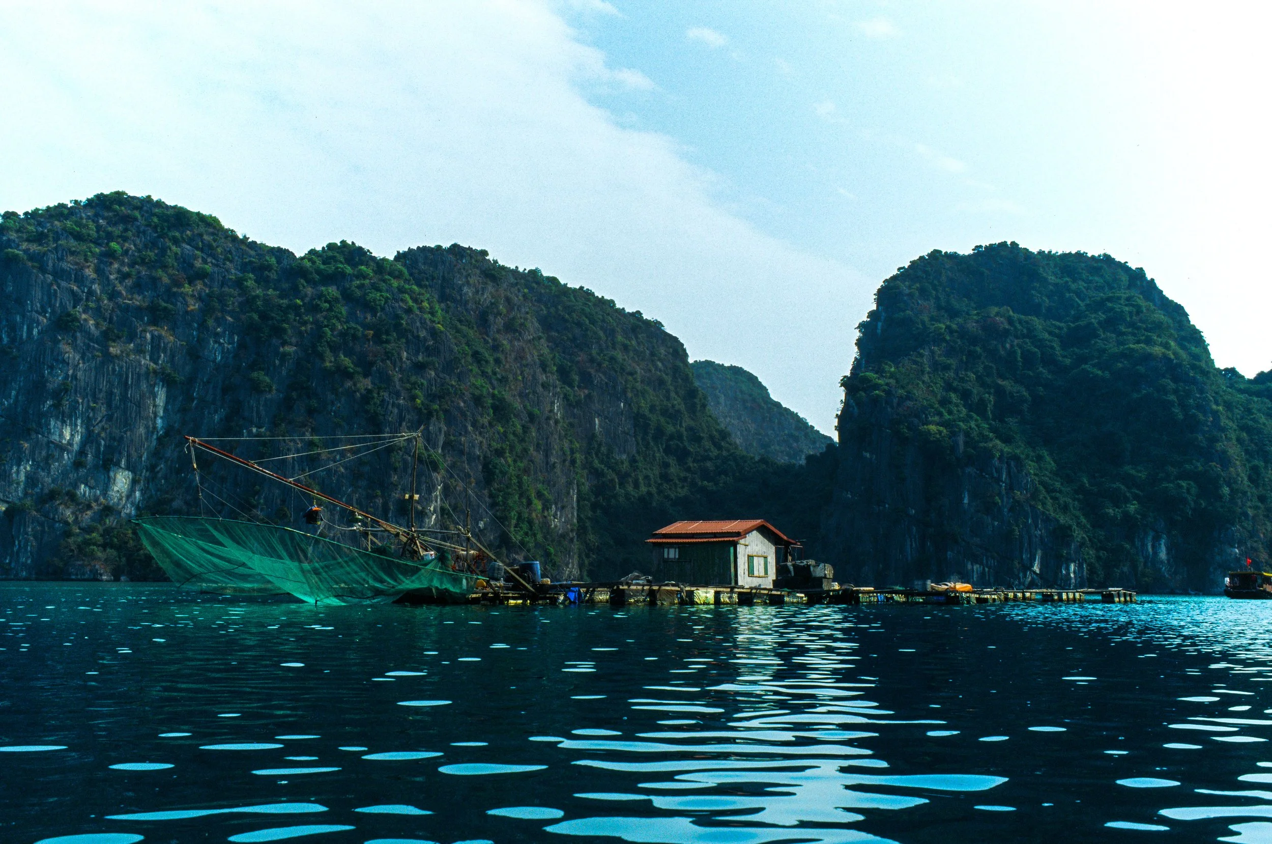 A body of water with ripples, a floating house with a rust-colored roof, and a fishing boat with a green net, set against limestone karst mountains and a partly cloudy sky.
