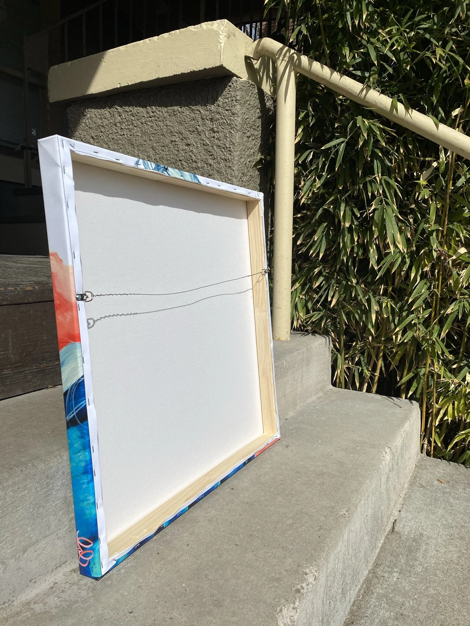 Back of a canvas painting with wire hanging, placed on outdoor concrete steps near bamboo plants and a handrail.