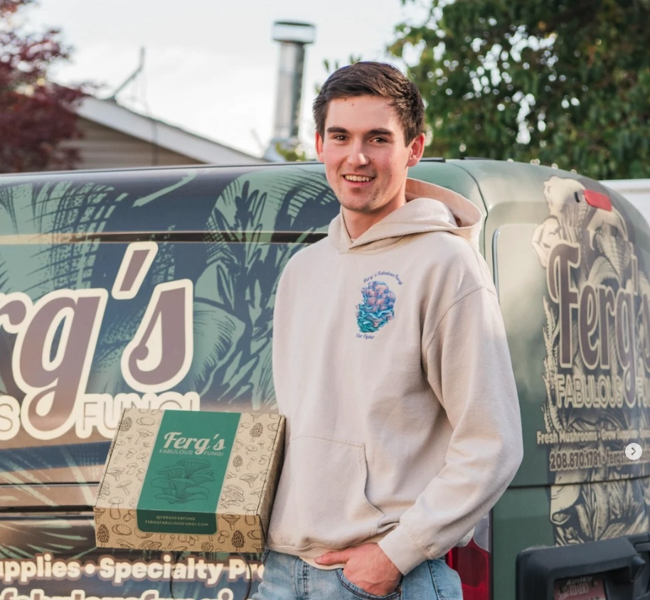 A young man with brown hair wearing a light-colored hoodie stands next to a van holding a cardboard box labeled 'Ferg's Fabulous Fungi.' The van has graphics and text promoting fresh mushrooms and a website.
