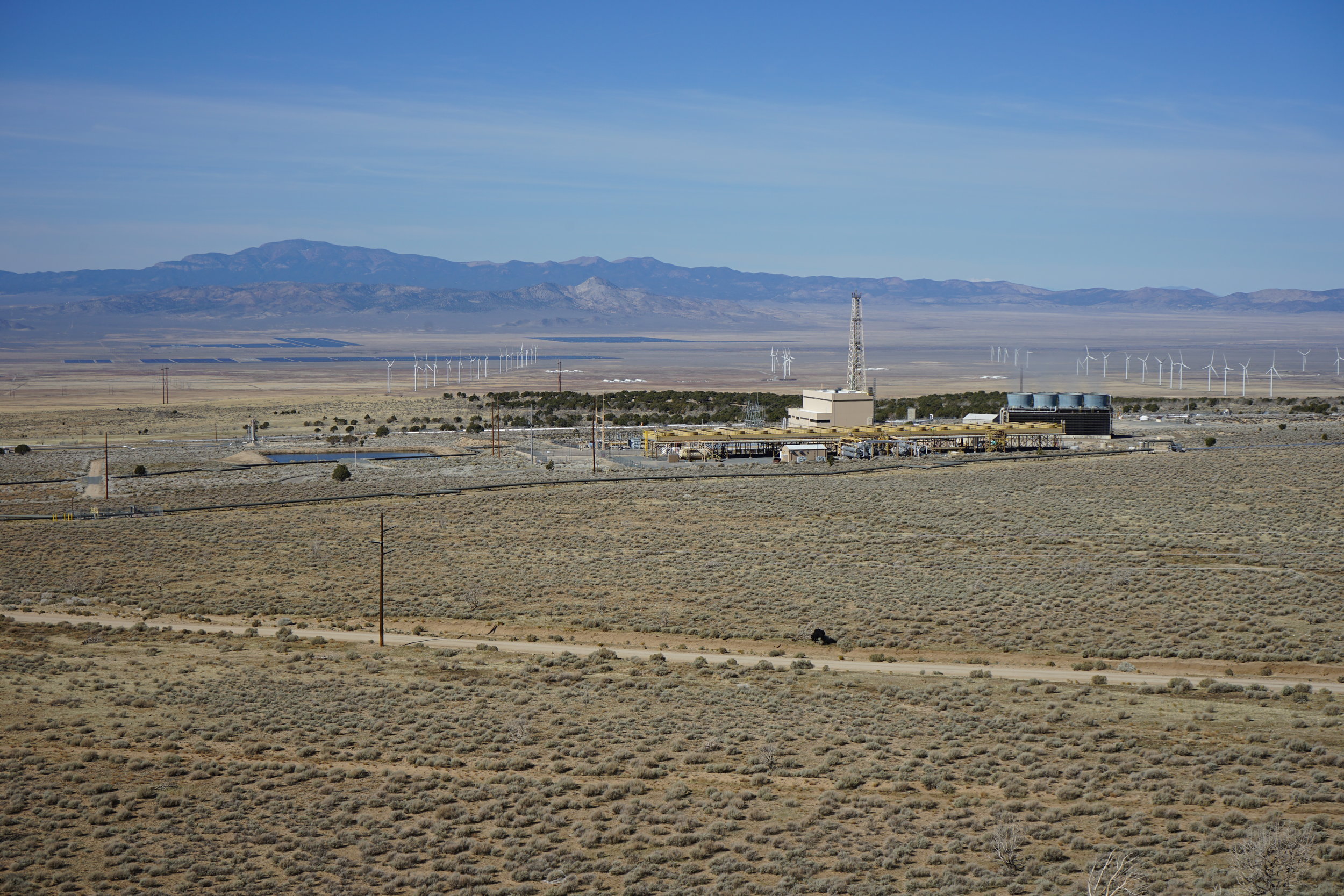The Milford Renewable Energy Corridor, looking WNW. In the foreground, the Blundell Geothermal Power Plant produces 34 MW of power from a hydrothermal system that appears to be bounded by a steep fault that roughly corresponds to the treed hillside behind the plant. The Milford FORGE site will be between the rows of wind turbines on the right side of the image. Across the valley, a large solar farm also produces electricity.