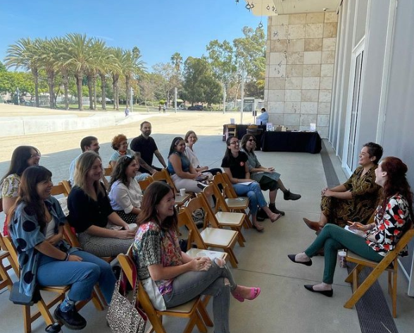 A group of people sitting on wooden chairs outdoors, participating in a discussion or presentation, with two women seated at the front facing the audience.