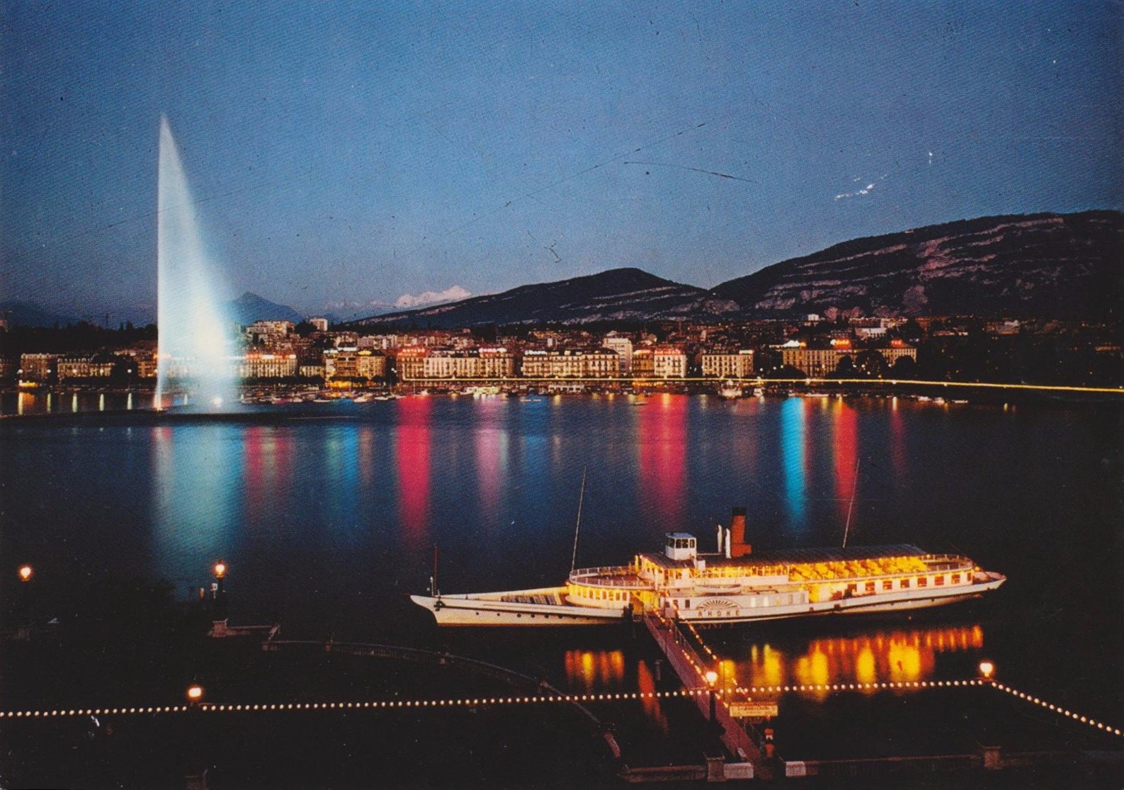 Vintage postcard of Geneva with a view of Lac Léman and a paddle boat, the Jet d’Eau, la Rade with colourful lights and the Salève.