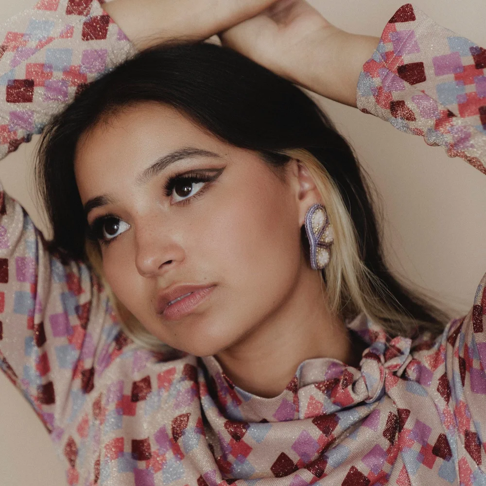 Kinsale Drake, a young Diné person with light brown skin and dark hair, looks off to the upper left as she raises her arms above her head. Behind her is a beige background.