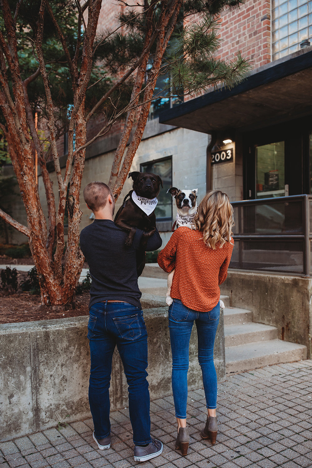 Engaged couple holding their dogs with backs faced towards camera. 