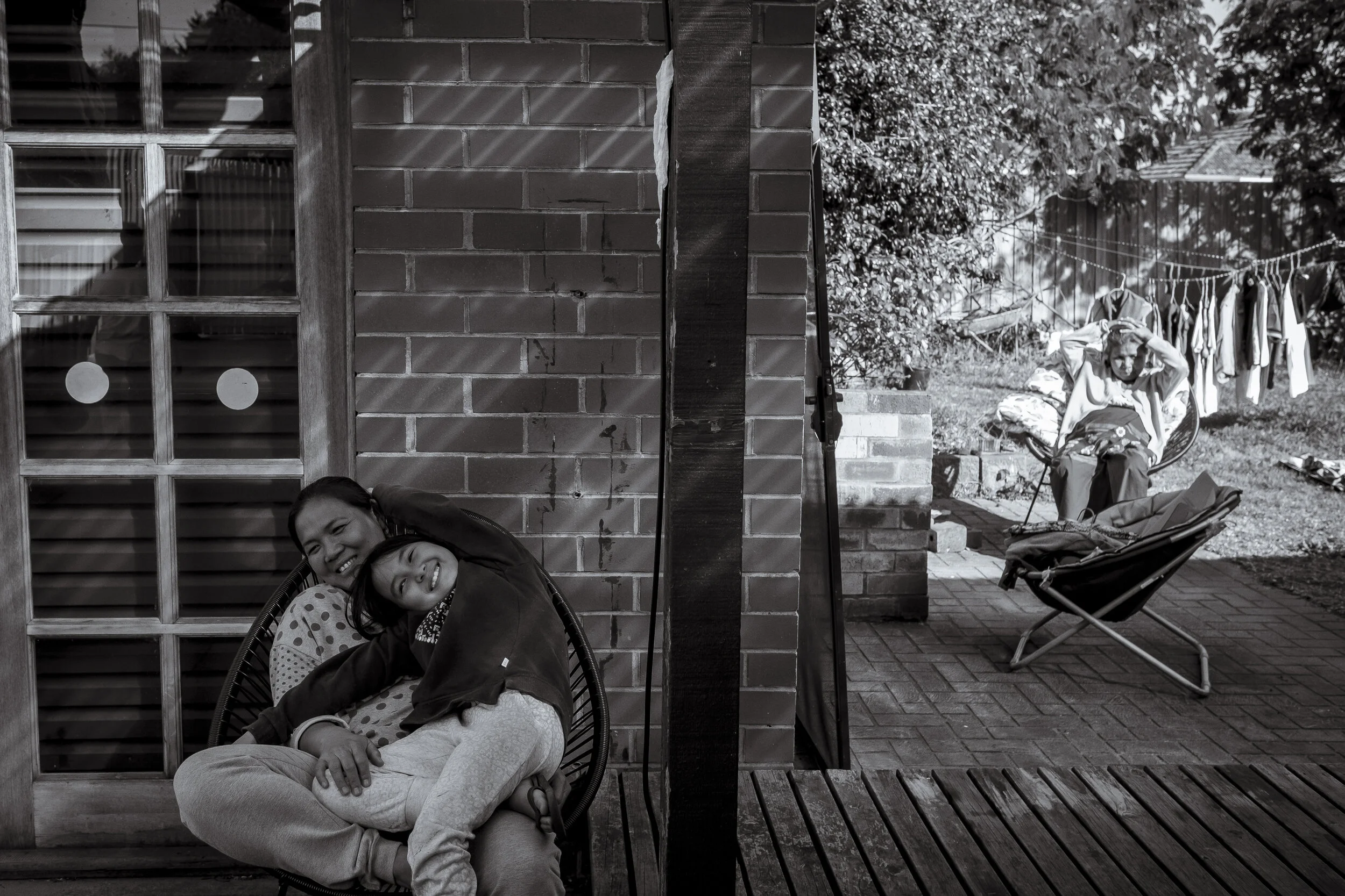  Wyndham, Melbourne. Ah Myang's daughter and grand-daughter share a lighter moment as Ah Myang's wife sits in the backyard alone. Elders will often find it the hardest amongst re-settled communities mainly due to their lack of, or ability to learn En