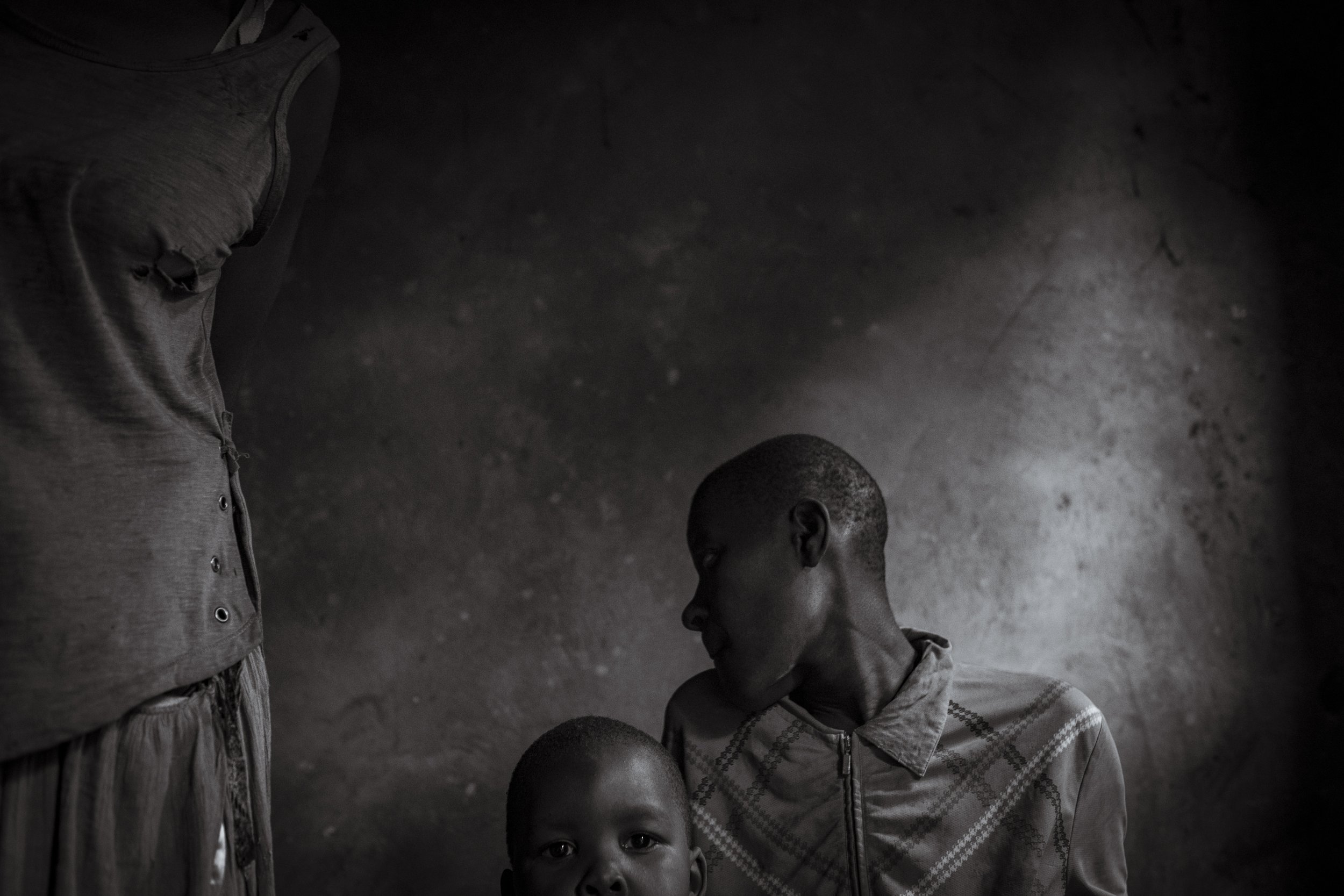  25 July 2023. Namazala, Jinja District, Uganda. Four-year-old Edwin Nangobi stares into the camera as he sits with his mother, 20-year-old Katherine Muwunguzi (r) and Grandmother Joy Nangobi  in the room Katherine shares as a sleeping quarter with b