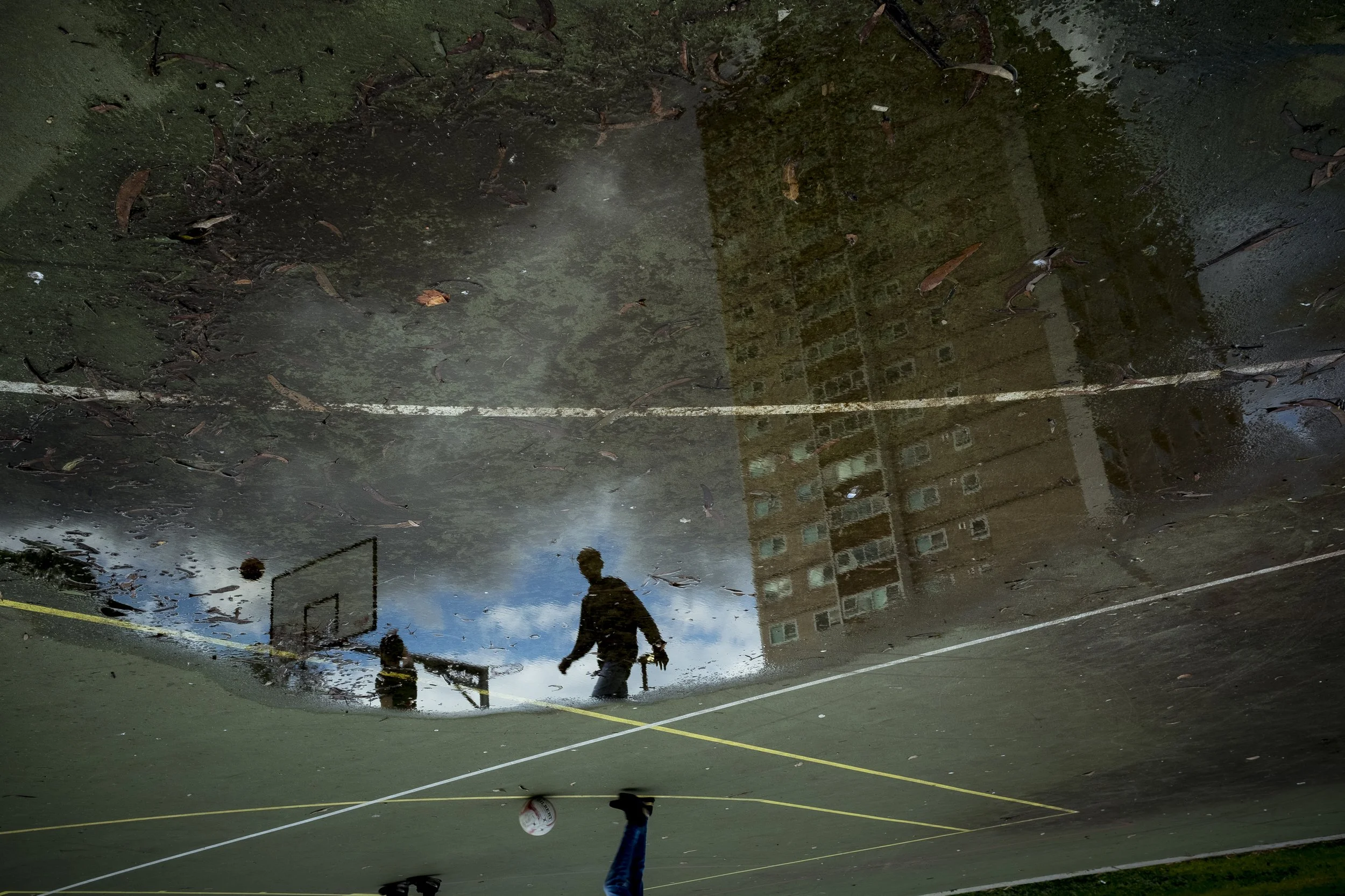  9 July 2020. Wellington Street, Collingwood, Australia. Residents playing basketball are dwarfed by the Collingwood public housing flats reflected in a puddle at Unity Park.  