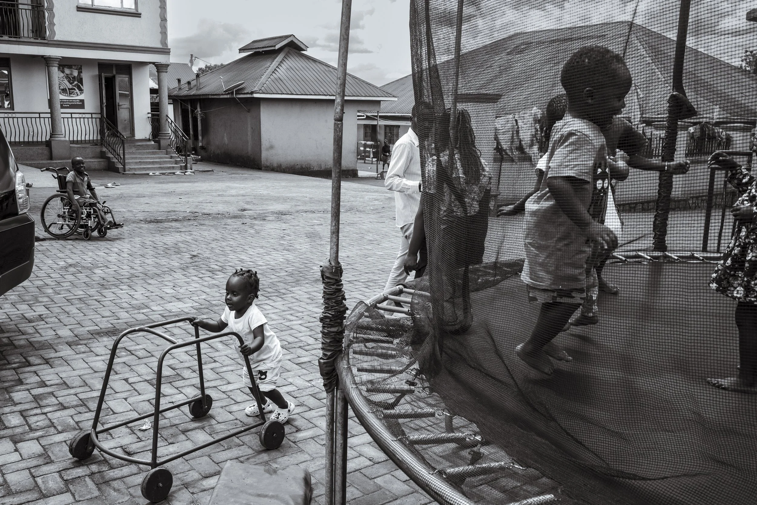  26 November 2024. Bugembe, Jinja District, Uganda.  As two-year-old Mildred, who lives with Down Syndrome, walks assisted by a walking frame, children of staff play unassisted on a trampoline at the Home of Hope. Every Tuesday is therapy day at the 
