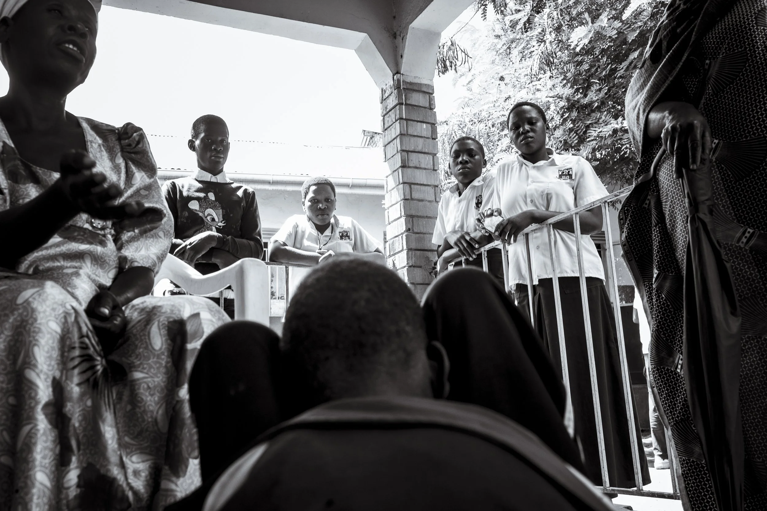  29 November 2024. Buwenge, Uganda. 16-year-old Moses is ogled by local students as his mother, Evelyn Mbwali explains his story to a health worker at a local outreach clinic.  Moses was brought to the outreach by his mother who was worried about his