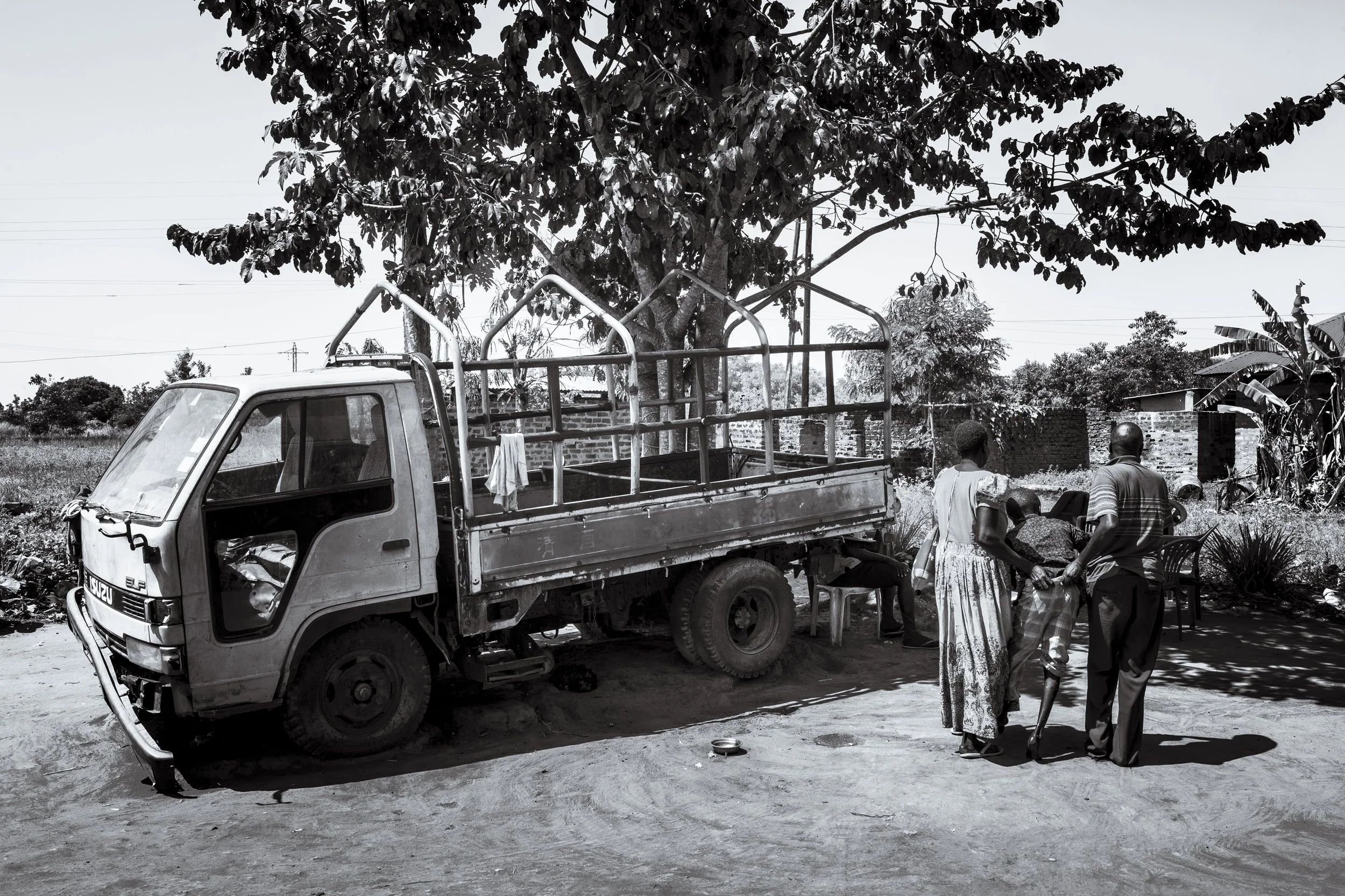  4 December 2024. Omalera, Soroti District, Uganda.  Local Witch doctor Robert Apedu and Florence Aguro carry Florence's son Noah Oyara into the shade after Robert treated Noah. Noah has no use of his legs and also lives with hydrocephalus. Robert tr