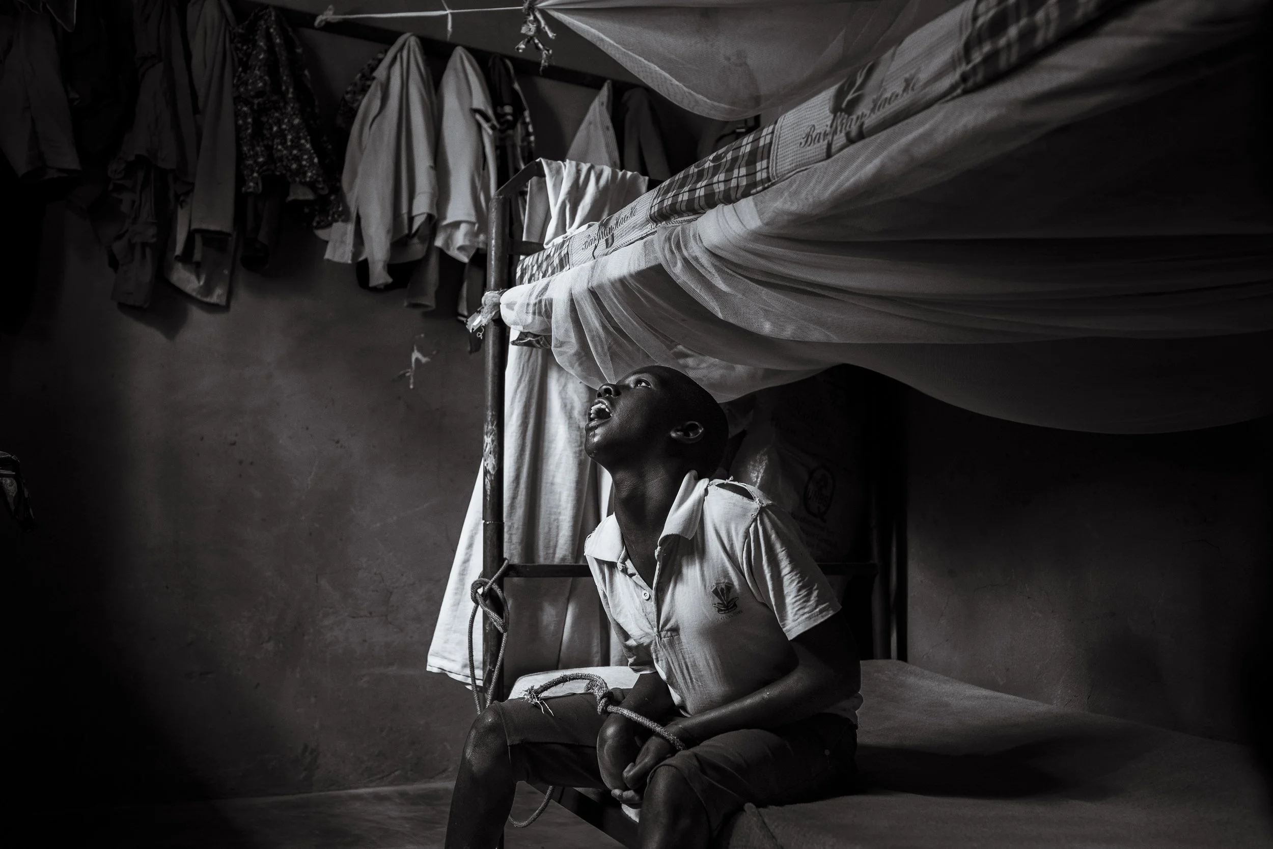  5 December 2024. Omalera, Soroti District, Uganda. 17 -year-old David Emwodu sits hands bound by a rope and tied to the uprights of the bunk bed he shares with his brother.   David lives, undiagnosed, with an intellectual disability which can see hi