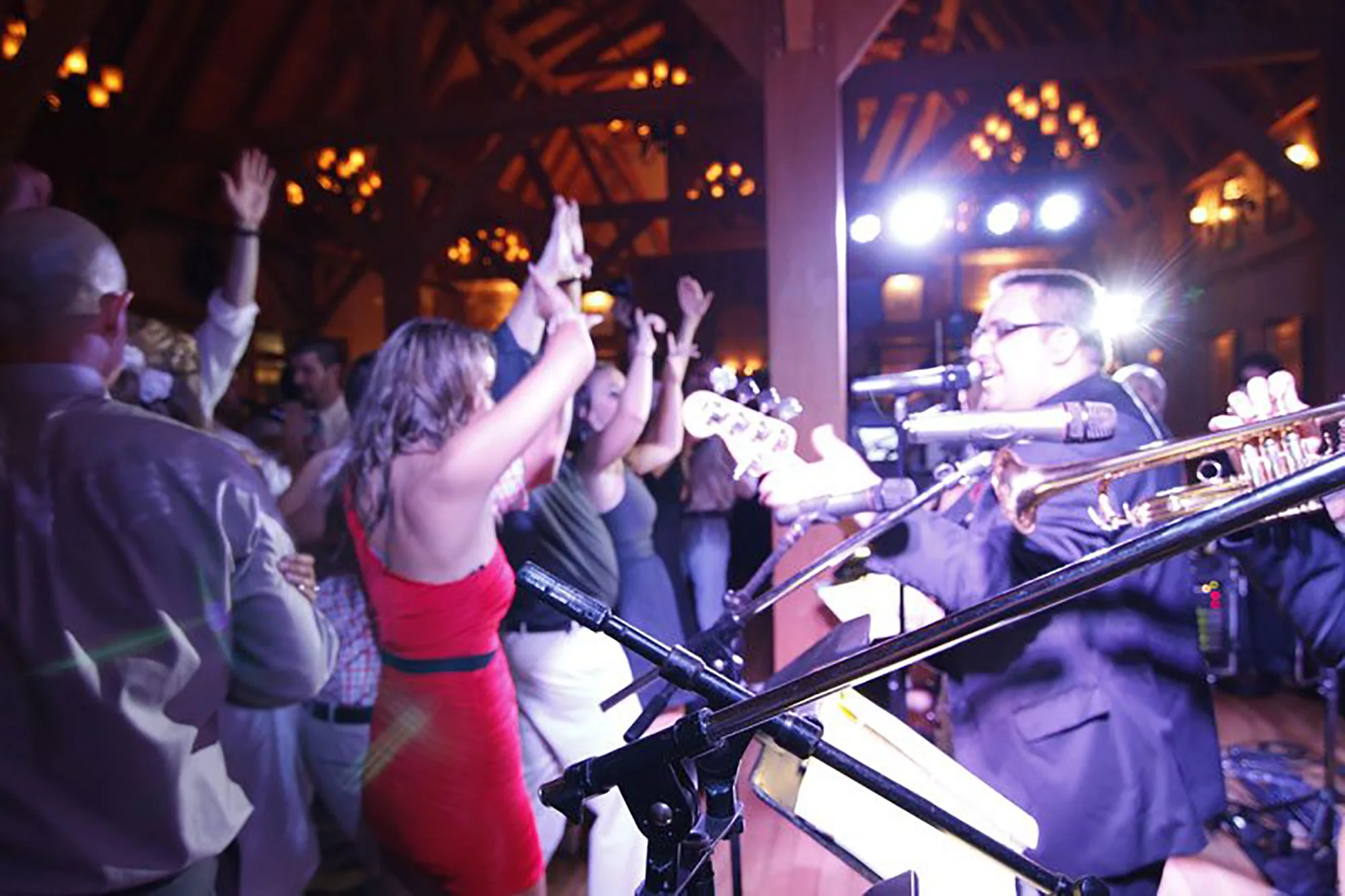 A band performing live at a concert with a crowd of people dancing and raising their hands in front of the stage. The band includes a singer and a musician playing a guitar or keyboard, with stage lights illuminating the scene.