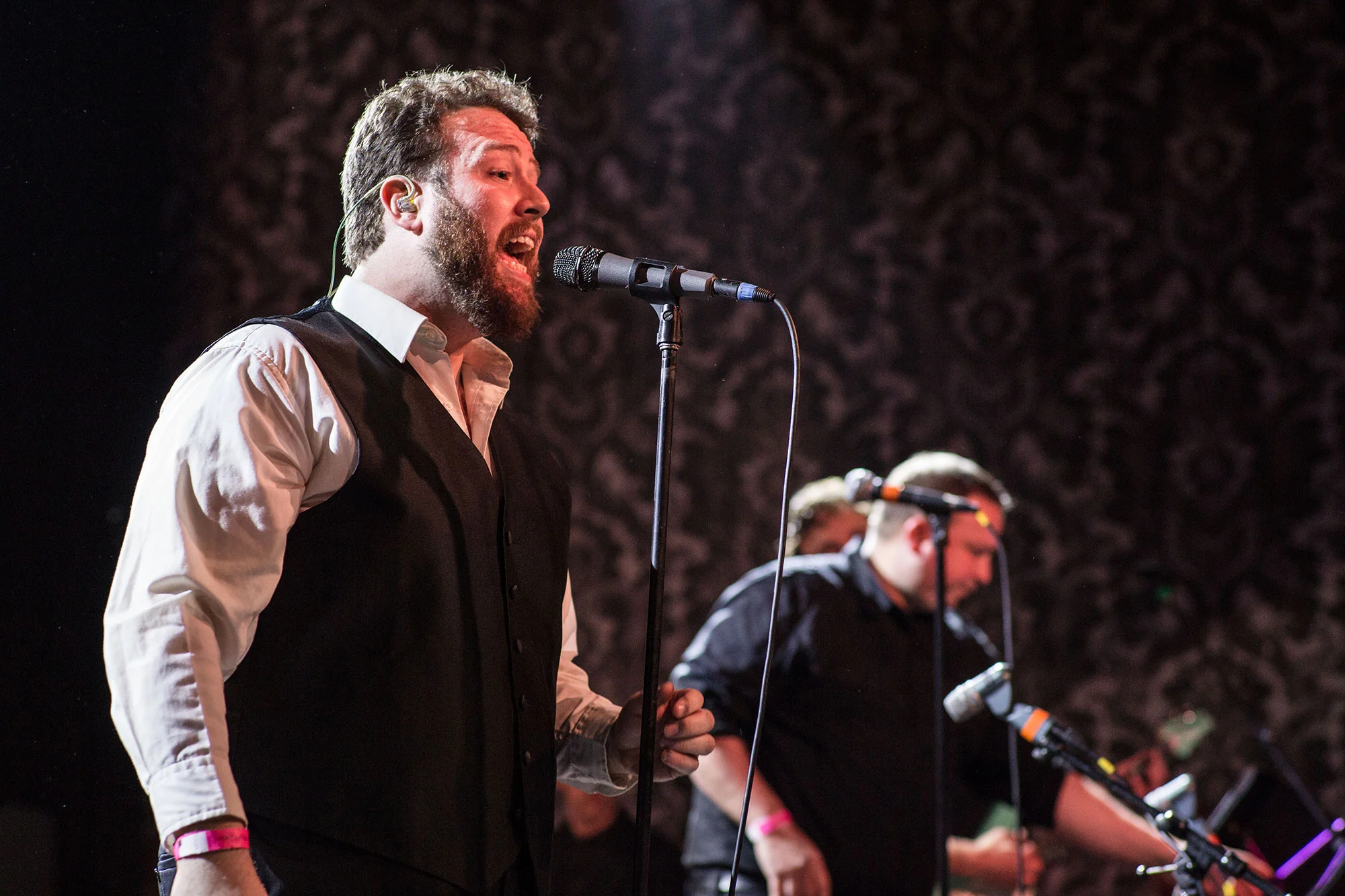 A male singer with a beard and wearing a white shirt and black vest performs into a microphone on stage. Two other musicians are in the background, also near microphones, with one looking at a device, against a dark patterned backdrop.