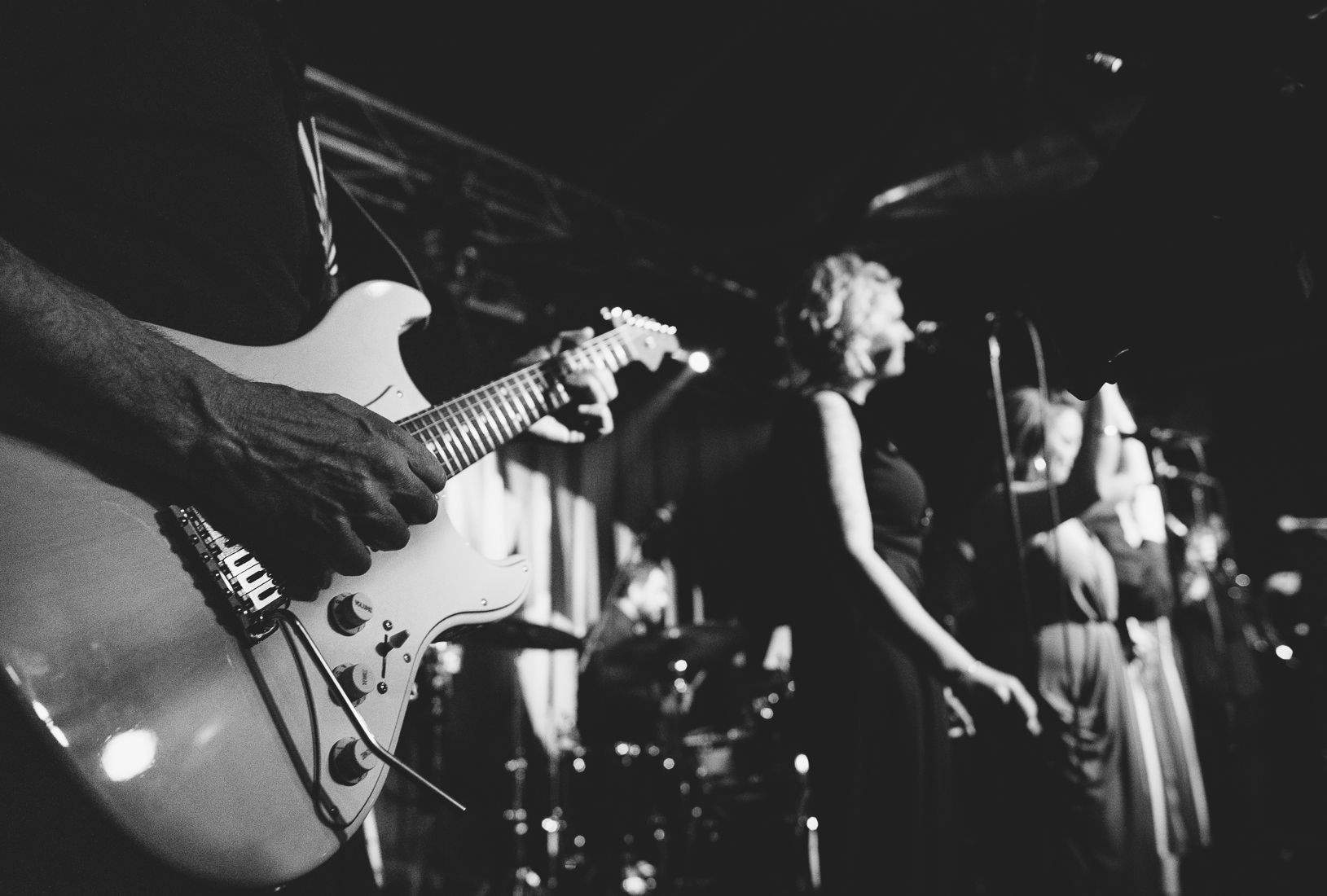Black and white photo of musicians performing on stage, with a focus on a guitarist in the foreground and a female singer in the background.