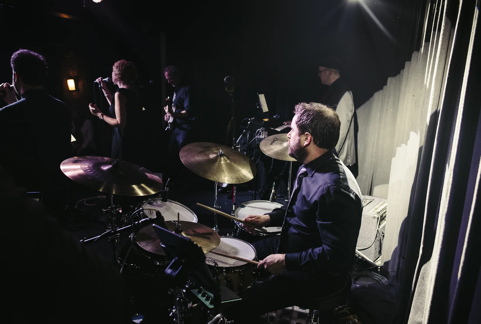A man playing drums on stage with nearby musicians and singers, dimly lit with stage lights.