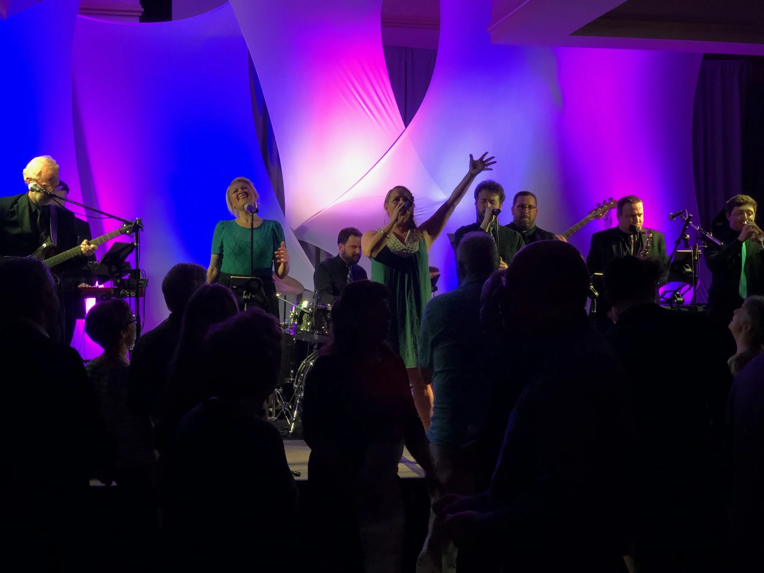 A live band performing on stage with colorful purple and blue lighting, with an audience watching in the foreground.