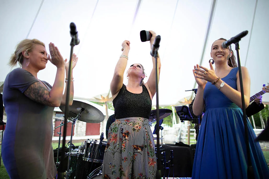 Three women on stage during a live music performance, with musical instruments and microphones, under a large tent with a bright background.