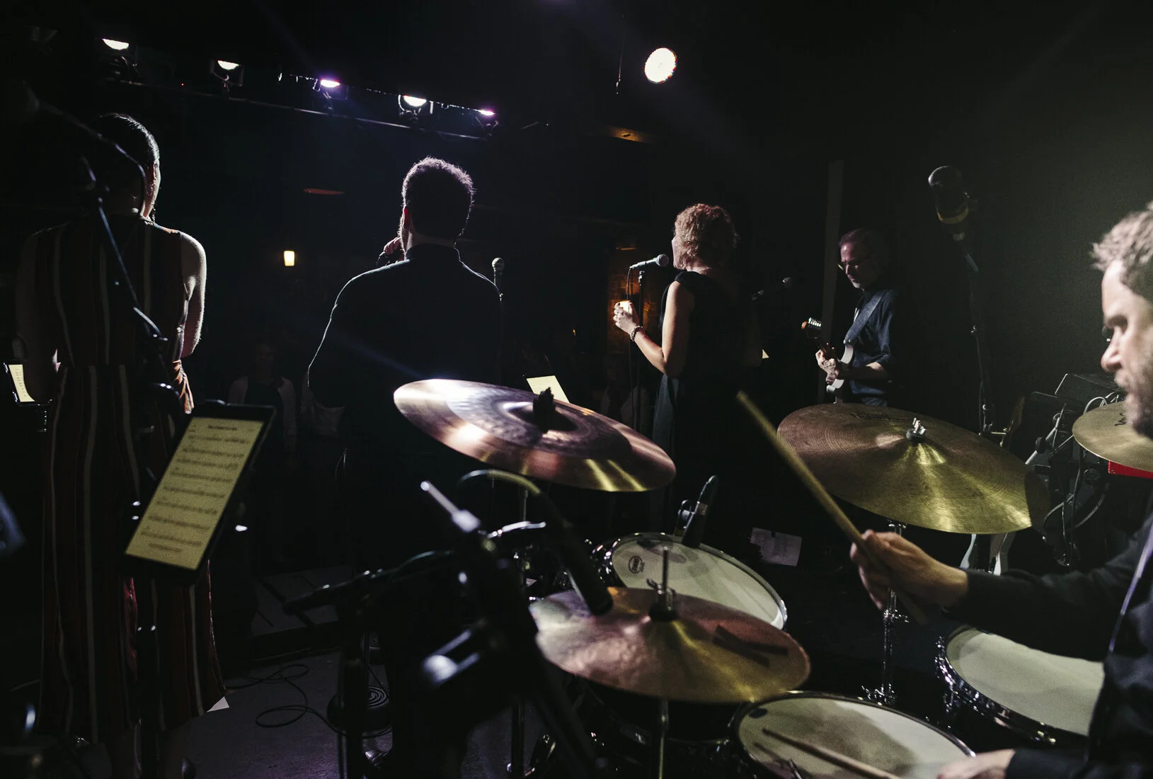 A live music performance on stage with musicians, including a drummer, a guitarist, and vocalists, illuminated by stage lighting.