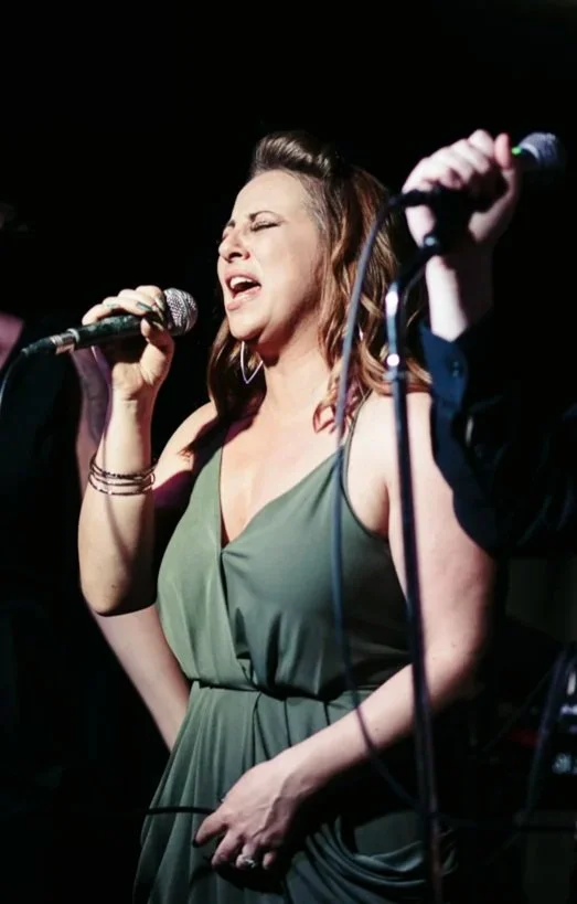 A woman singing into a microphone during a live performance, wearing a green dress and earrings with a passionate expression.