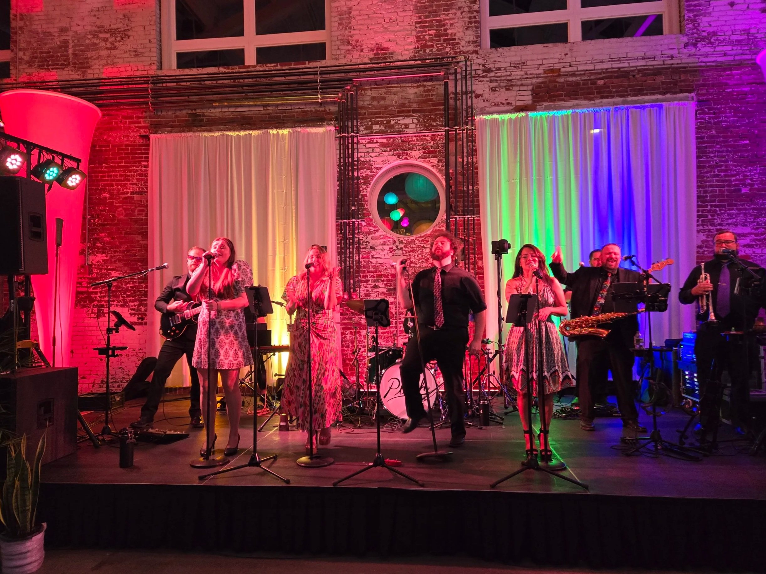 A multi-piece band performs on a stage with colorful lighting, including pink, yellow, and green, in front of a brick wall with white curtains and a circular window.