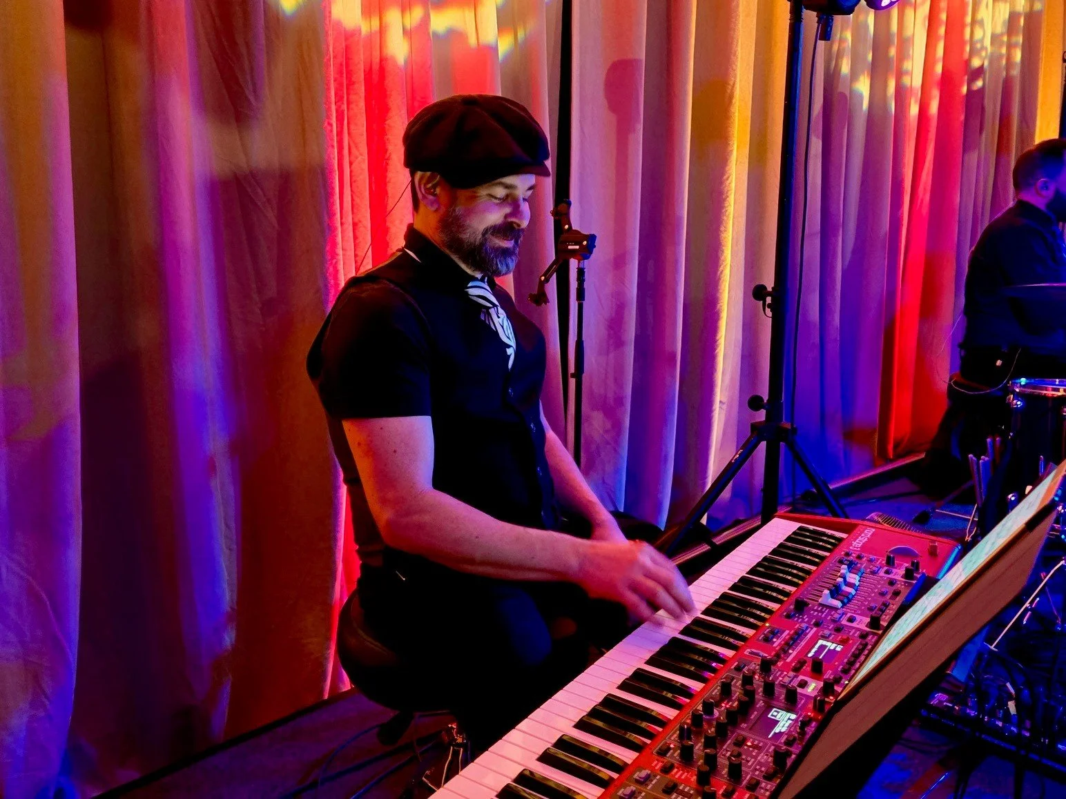 A man wearing a black beret and black shirt playing an electronic keyboard on stage, with multicolored stage curtains behind him.
