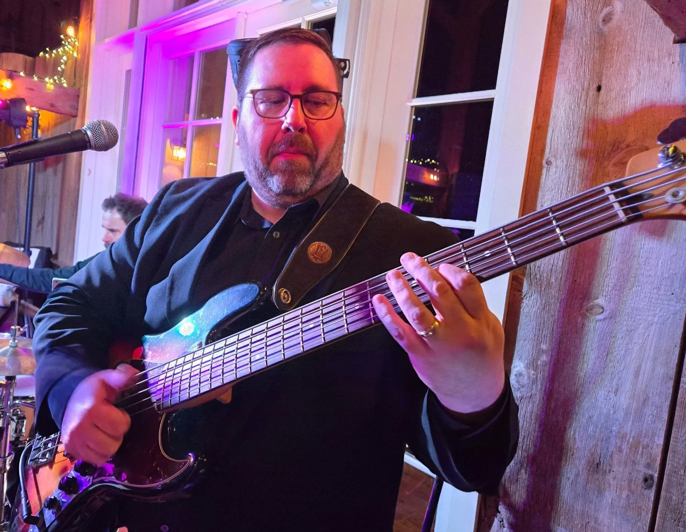 A man wearing glasses and a black shirt playing an electric guitar at a cozy indoor event with colorful ambient lighting.