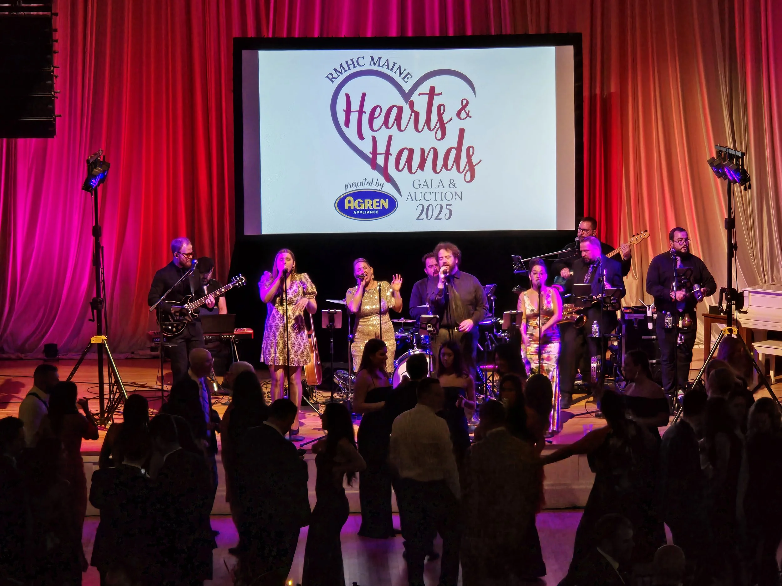Live band performing on stage at Hearts & Hands Gala & Auction 2025 with a large screen displaying the event logo, surrounded by red and gold curtains, and an audience dancing in front.