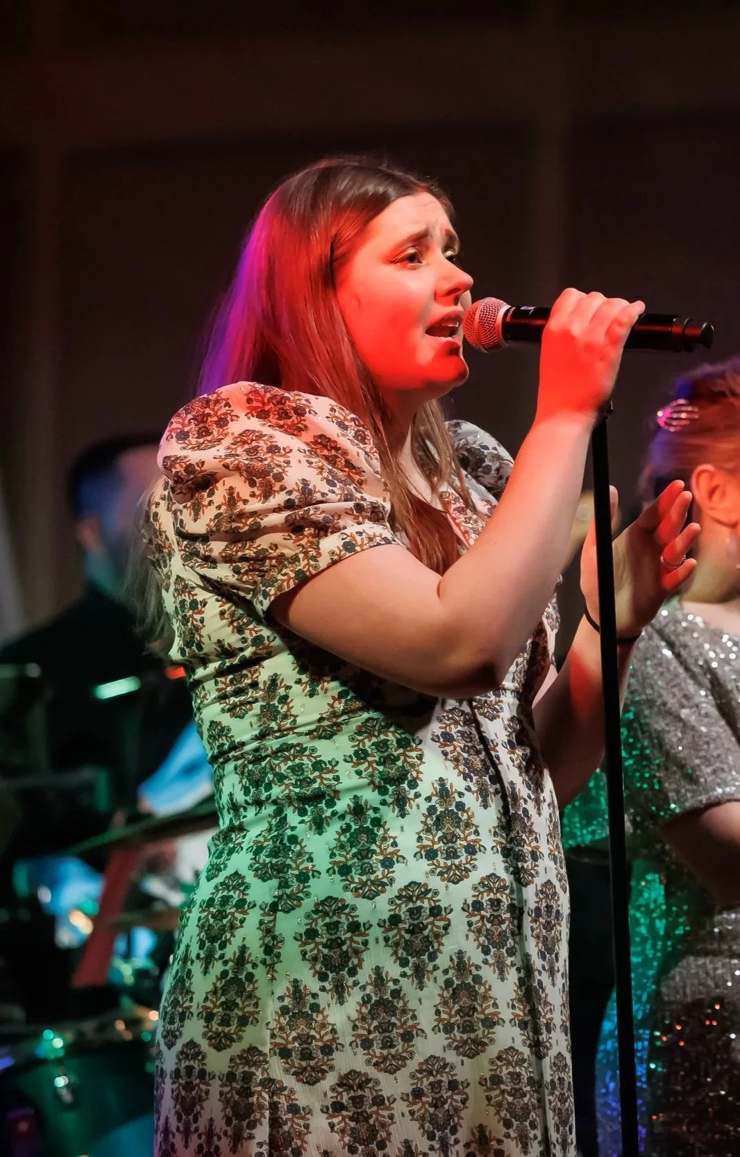 A young woman singing into a microphone on stage, wearing a floral patterned dress, with colorful stage lighting.