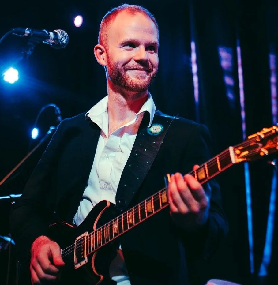 A man with a short beard and mustache playing an electric guitar on stage, wearing a black suit and white shirt, with stage lights and a microphone in the background.