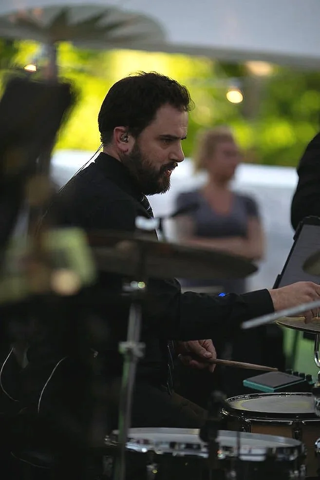 A man with dark hair and a beard playing drums at an outdoor event, with a woman blurred in the background.
