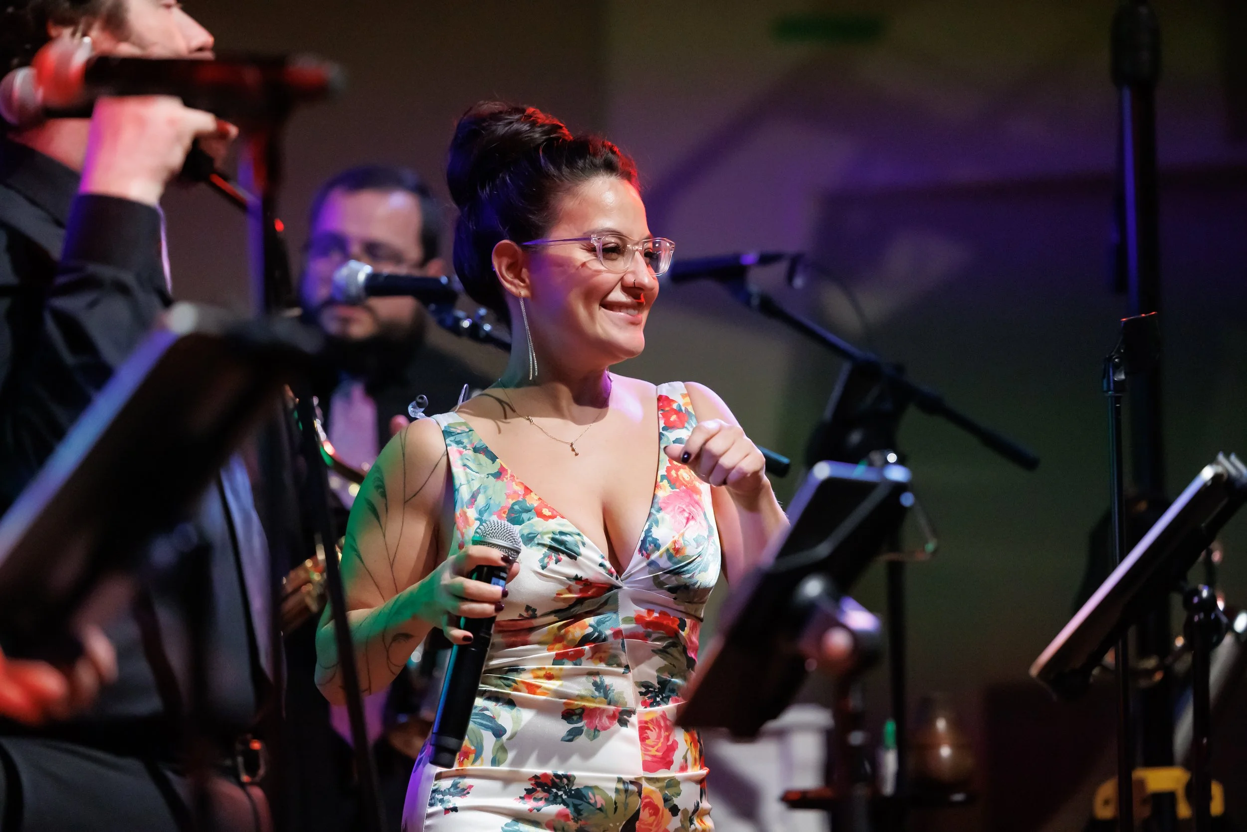 A woman in a floral dress holding a microphone, smiling, with musicians playing instruments around her in a concert setting