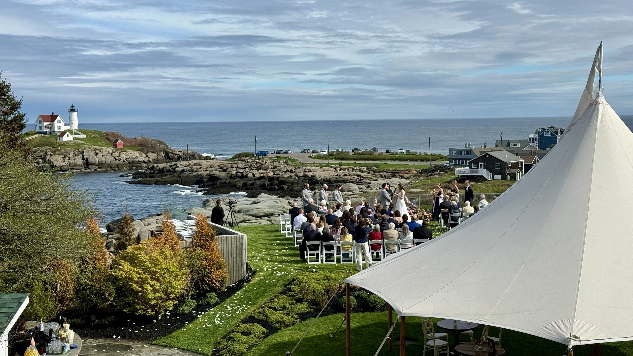 Outdoor wedding ceremony near a rocky coastline with a lighthouse in the distance. Guests seated in chairs, the bride and groom at the altar, and a large white tent in the foreground.
