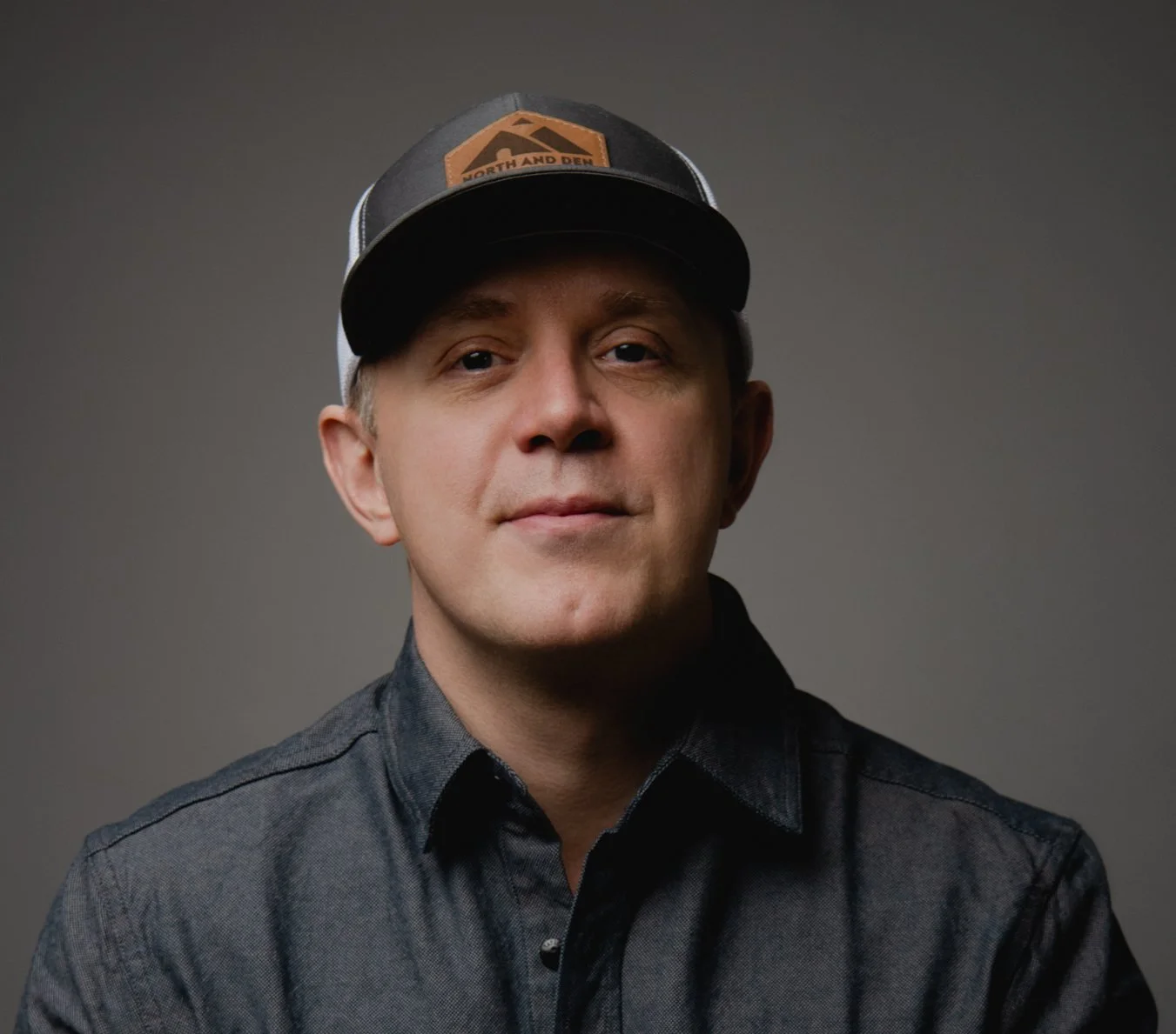 Patrick Ermlich head of Compass Method Consulting wearing a dark button-up shirt and a trucker cap with a patch reading 'North and Den' and a mountain logo.