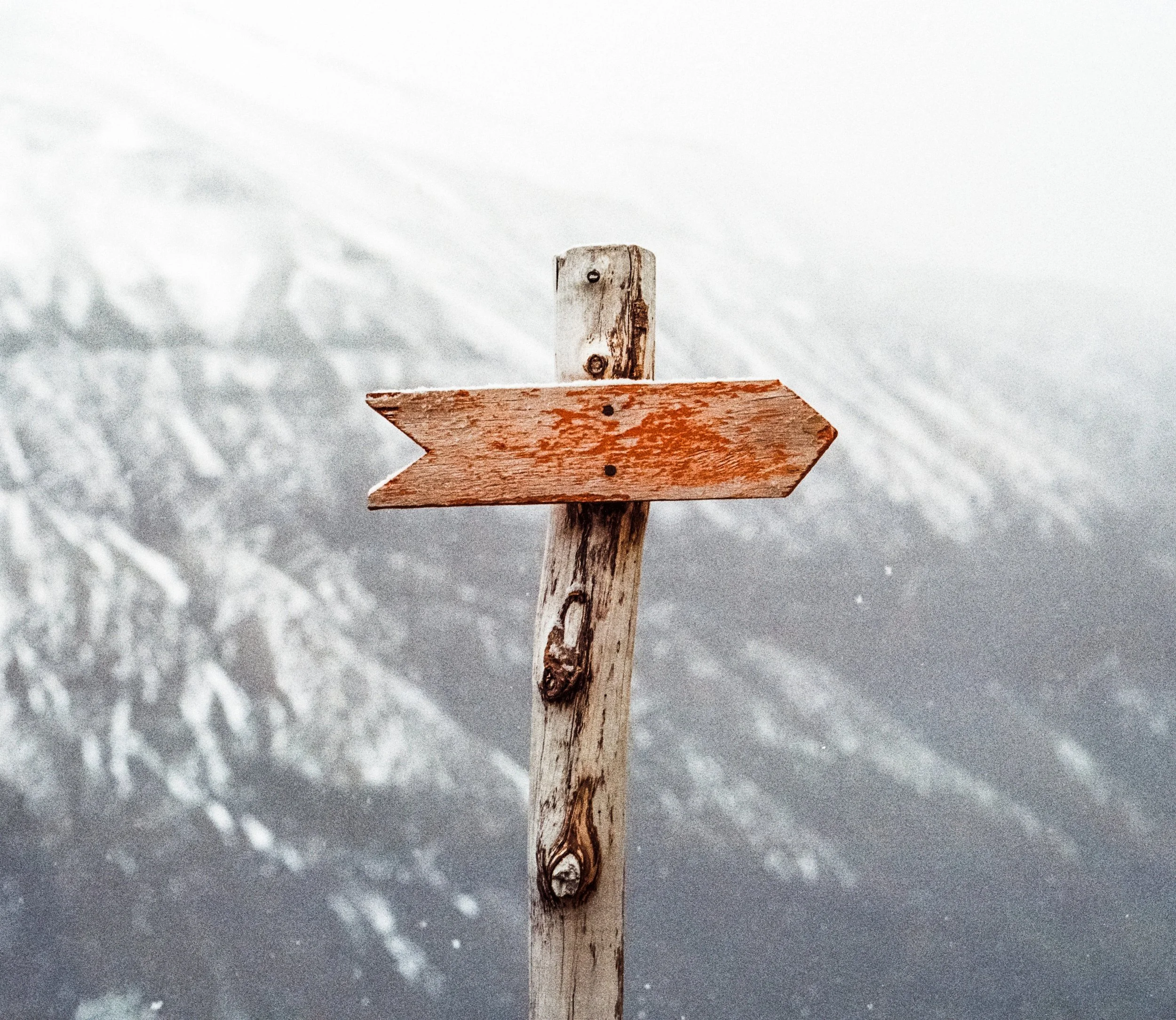 A weathered wooden signpost with an arrow, set against a snowy mountain background.