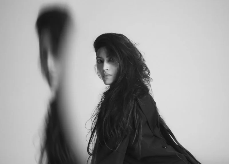 music artist Vandana with long, wavy hair looking over her shoulder at the camera, with a blurred figure in the foreground, all in black and white.