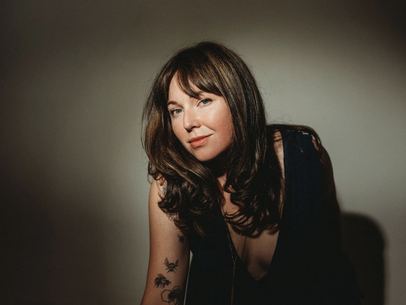 artist coach Holly Pyle with long brown hair, wearing a black top, poses against a plain neutral background, looking at the camera with a slight smile.