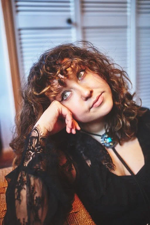 Artist Ellie Fern with curly hair resting her head on her hand, looking thoughtfully to the side.