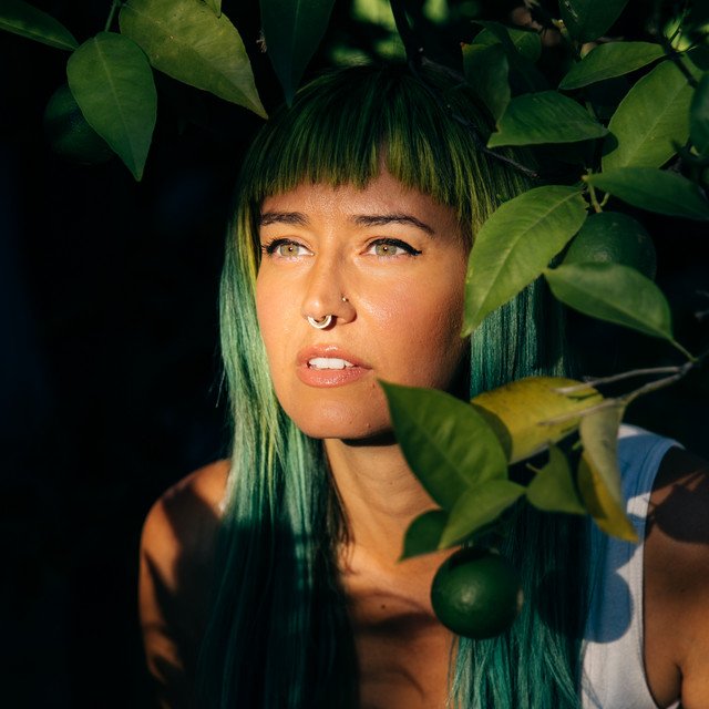 artist coach Lila Rose with teal hair and septum piercing among green leaves and a lemon hanging from a branch.