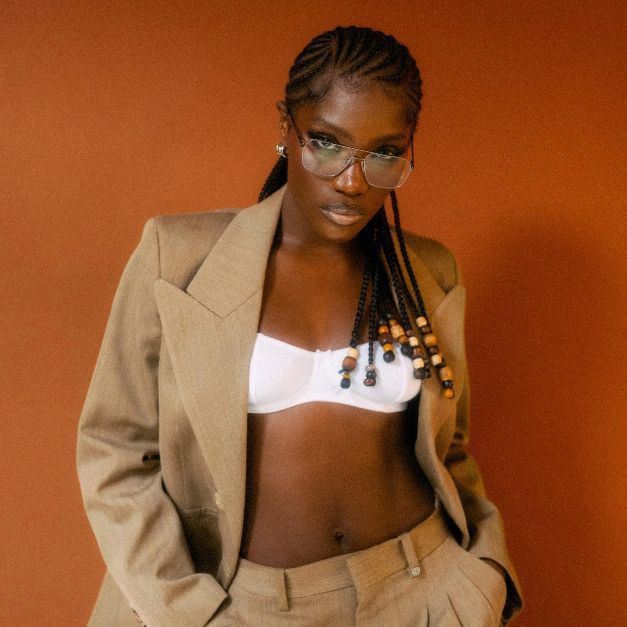 the artist doechii wearing glasses, a beige blazer, and matching beige pants, with a white bandeau top, stands against a warm brown background.
