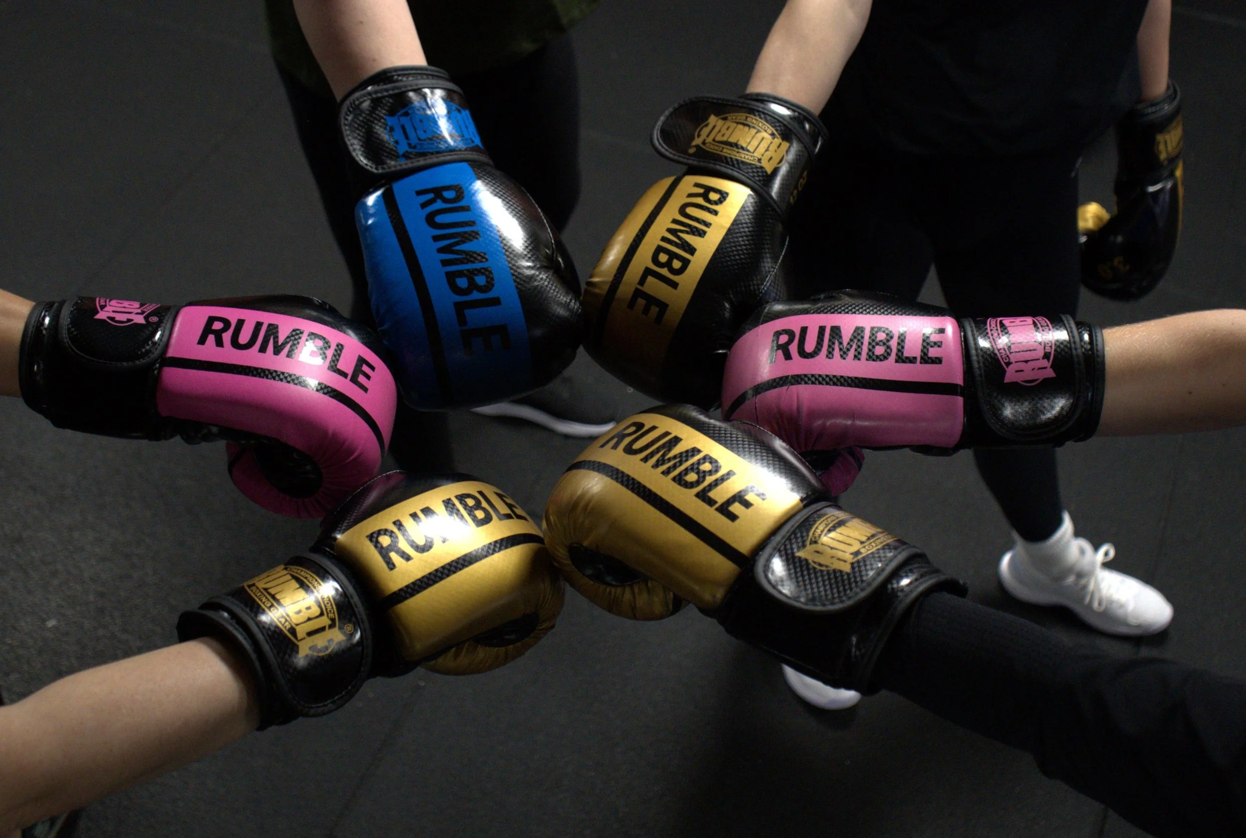Zes handen in de boksring die verschillende kleuren bokshandschoenen met de tekst 'RUMBLE' en 'RUBBLE' omhoog laten zien, in een cirkelvorm, op een donkere vloer.