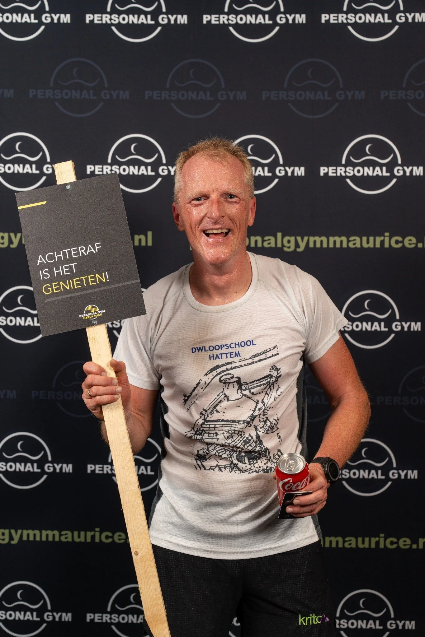 Een man die glimlacht, die in een sportschoolpose voor een achtergrond met het logo van Personal Gym staat, houdt een bordje met de tekst 'Achteraf is het genieten!' en een blikje Coca-Cola in zijn andere hand.