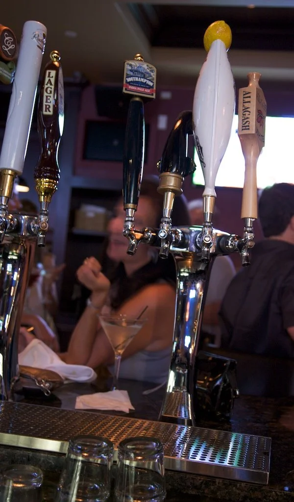 Four beer tap handles on a bar, with a woman and a man in the background.