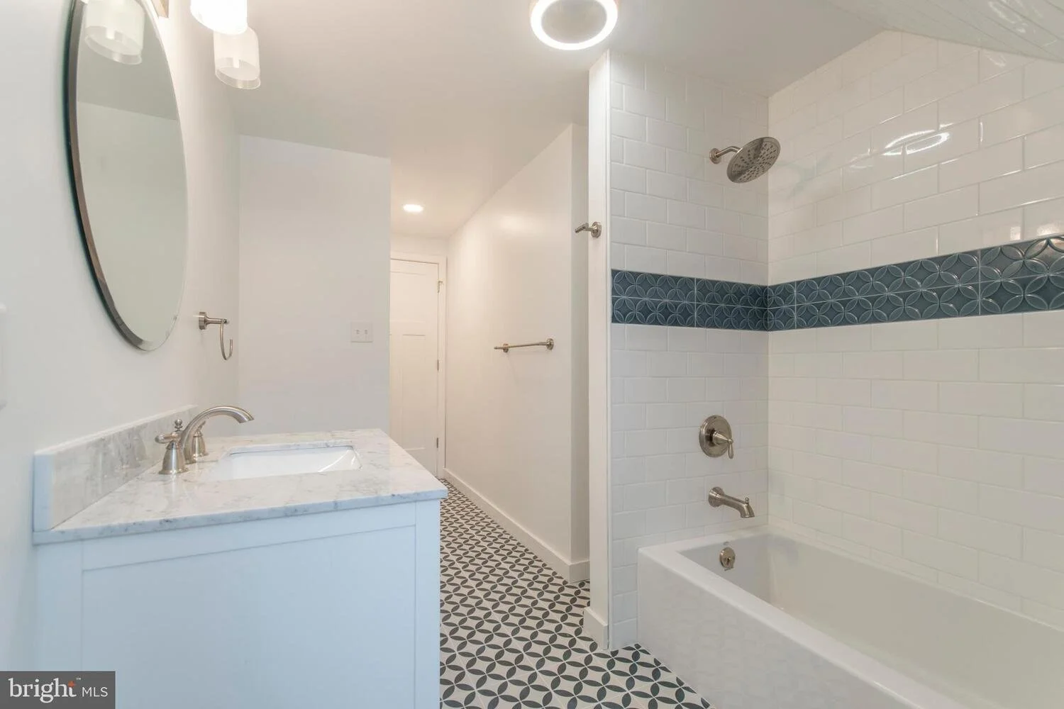 Bathroom with a white vanity and marble countertop, an oval mirror, and a shower with white brick tiles and blue decorative band, a bathtub with multiple water fixtures, and black and white patterned floor tiles.