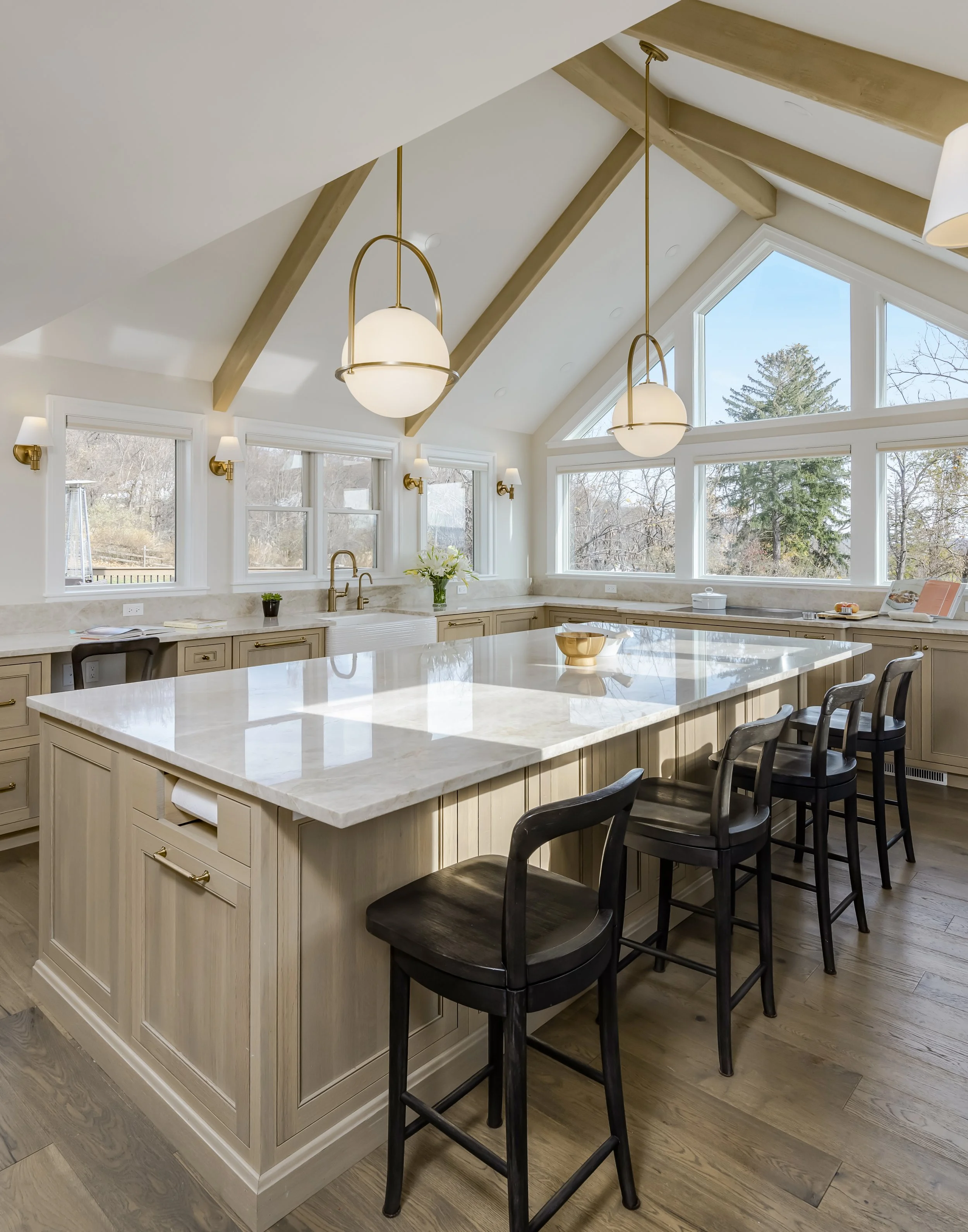 Modern kitchen with large white marble island, black chairs, multiple windows, and two pendant lights hanging from the ceiling.