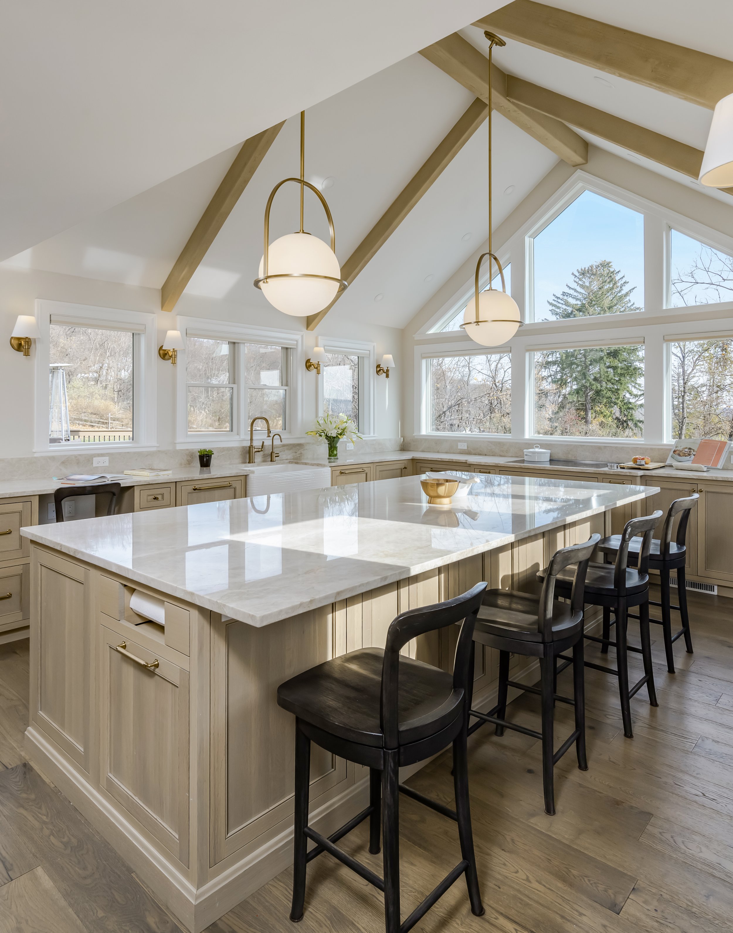 Modern kitchen with beige cabinets, a large white marble island, black bar stools, a window with a view of trees, and pendant lights hanging from a sloped ceiling with wooden beams.